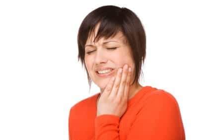 Woman in orange shirt holding cheek, smiling with eyes closed, suggesting dental pain or discomfort.