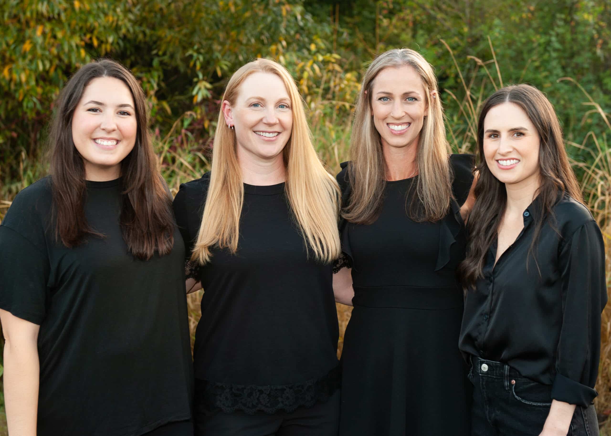 Four female dental professionals smiling at camera outdoors with green foliage background.