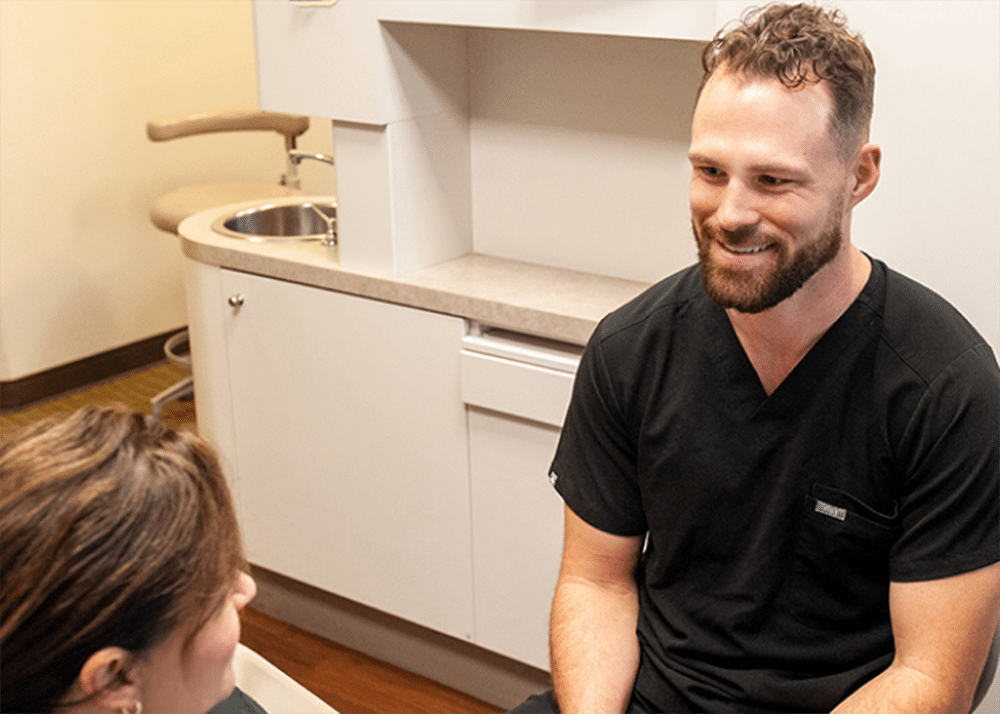 Male dentist in black scrubs smiling while consulting with female patient in dental office.