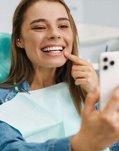 Young woman in dental bib smiling and pointing to teeth in dental office setting.