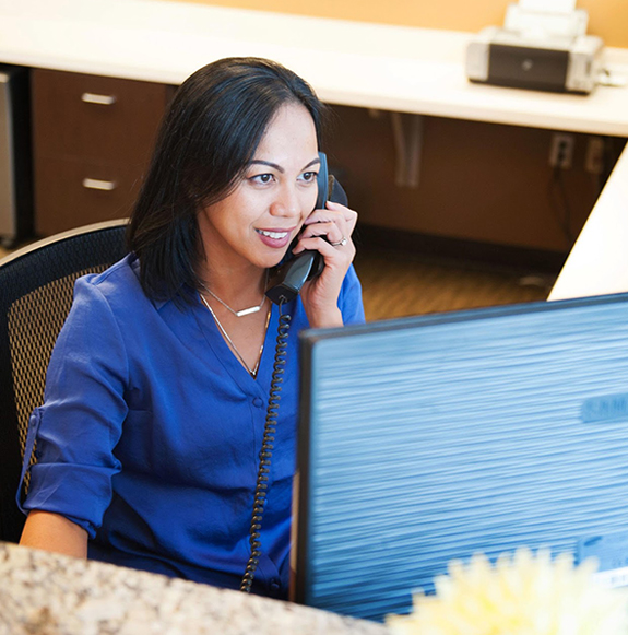 Female dental receptionist in blue scrubs speaking on phone at office desk.