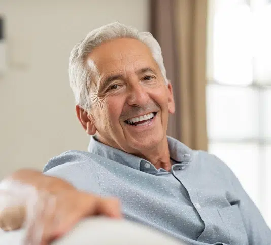 Smiling older man in light blue shirt sitting indoors by window at dental office.