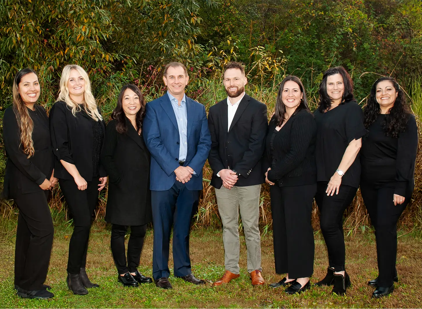 Professional dental team of nine people posing together outdoors against green foliage background.